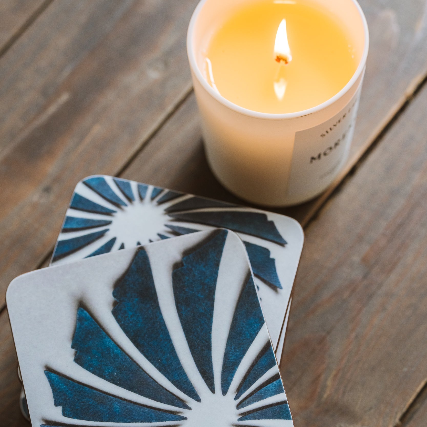 Coaster with blue and white design on a wooden surface with a lit candle.